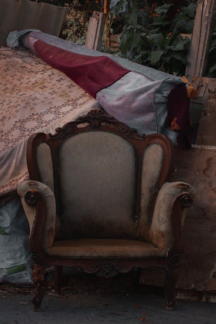 In the foreground, there is an antique upholstered armchair with a wooden frame featuring ornate carvings. The upholstery is a faded beige fabric, and the armrests are curved with detailed woodwork. Positioned behind the armchair, a rolled-up piece of fabric or possibly a blanket with grey, maroon, and dark grey sections is draped over a surface, partially covering it. The background shows a cluttered outdoor or semi-indoor space with wooden slats, green foliage, and additional miscellaneous items that suggest a storage or clearance area. The lighting appears natural, with a dim ambiance, indicating an outdoor setting. This scene relates to furniture or household item clearance, possibly handled by a private rubbish removal service such as Rubbish Clearance Bromley, emphasizing the context of alternative waste handling or on-site clearance of bulky items.