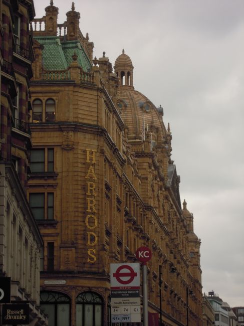 The image depicts an ornate, historic building with a detailed façade constructed from yellowish sandstone, featuring decorative arches, columns, and intricate stonework. The building's upper levels are crowned with a large dome, smaller domes, and spire-like structures, showcasing elaborate architectural design typical of a historic city centre. Adjacent to the building, a street sign indicates a London Underground station for South Kensington, with a circular red and white London Underground symbol and a bus route information sign beneath. The scene is set outdoors under overcast skies, with ambient natural lighting casting subtle shadows on the building's textured surface. The foreground includes part of a pavement and street level, emphasizing the urban environment, which is characteristic of central London streets where private waste clearance services, such as those offered by Rubbish Clearance Bromley, often operate to manage rubbish and unwanted items from nearby residential or commercial properties, in contrast to council-led waste collection methods. This scene subtly highlights the importance of professional rubbish removal in historic urban settings where on-site clearance of accumulated waste and furniture is sometimes required to maintain the area’s aesthetic and cleanliness.