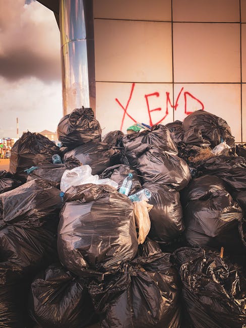 A large pile of black rubbish bags stacked against a building wall with orange graffiti text 'XEND' spray-painted on beige tiled surface. The bags are made of plastic, appear full and tightly tied, and some have visible creases and wrinkles. Among the bags, a few discarded plastic bottles are noticeable, with transparent and blue hues. The scene is outdoors, possibly on a city street or alleyway, with part of a metallic structure or column visible on the left side and cloudy skies overhead. The overall environment suggests an area awaiting rubbish collection or disposal, emphasizing the importance of alternative waste handling and private rubbish clearance services provided by Rubbish Clearance Bromley. The photographic lighting highlights the contrast between the dark rubbish bags and the lighter tiled background, capturing the clutter and disorder typical of unmanaged waste accumulation in urban settings.