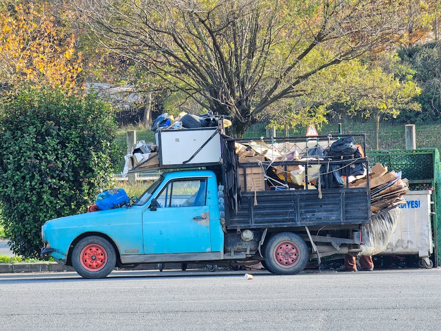 A small, old blue pickup truck with a black cargo bed is parked on the side of a street during daytime. The vehicle's exterior shows signs of wear, with faded paint and visible rust on the red-painted wheels. The truck is loaded with a large amount of mixed rubbish, including cardboard boxes, black plastic garbage bags, and miscellaneous debris, some of which are loosely stacked and spilling over the sides. The load extends above the height of the truck's sides, secured loosely with ropes. In the background, there is a tree with sparse green and yellow leaves, indicating early autumn, and a green fence lining the edge of a park or yard, with additional foliage visible further back. The scene is lit by natural daylight, with soft shadows cast on the pavement, capturing an instance of private waste disposal or alternative rubbish collection managed by local operators such as Rubbish Clearance Bromley.