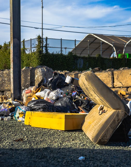 A large pile of black rubbish bags stacked against a building wall with orange graffiti text 'XEND' spray-painted on beige tiled surface. The bags are made of plastic, appear full and tightly tied, and some have visible creases and wrinkles. Among the bags, a few discarded plastic bottles are noticeable, with transparent and blue hues. The scene is outdoors, possibly on a city street or alleyway, with part of a metallic structure or column visible on the left side and cloudy skies overhead. The overall environment suggests an area awaiting rubbish collection or disposal, emphasizing the importance of alternative waste handling and private rubbish clearance services provided by Rubbish Clearance Bromley. The photographic lighting highlights the contrast between the dark rubbish bags and the lighter tiled background, capturing the clutter and disorder typical of unmanaged waste accumulation in urban settings.