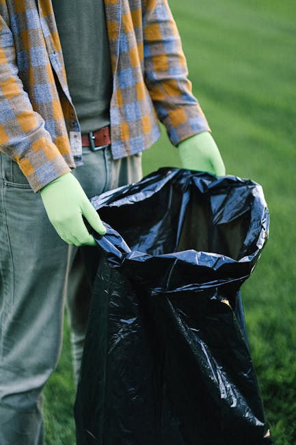 A person wearing a plaid shirt with shades of brown, yellow, and gray, along with a green t-shirt underneath, is holding open a large black plastic rubbish bag outdoors on a grassy area. The individual is wearing light green gloves, suggesting they are engaged in waste collection or rubbish removal. The scene shows the person's torso and hands, with a background of well-maintained green grass, indicating an outdoor environment such as a garden or park. The black rubbish bag appears to be partially filled or ready for collection, and the person is preparing it for disposal, possibly as part of a private waste clearance service provided by Rubbish Clearance Bromley, aligned with the theme of on-site rubbish removal or alternative waste handling solutions. The lighting is natural, emphasizing the textures of the plastic bag and the clothing, with no other objects or waste visible in the frame.